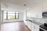 This single-floor unit showcases a clean, contemporary design with an open-concept layout that seamlessly connects the kitchen and living areas. The kitchen is well-appointed with floor-to-ceiling white cabinetry featuring brushed nickel hardware, gray quartz countertops, and a light gray subway tile backsplash. Stainless steel appliances include a refrigerator, electric range, built-in microwave, and dishwasher, providing a fully equipped cooking space. Warm-toned hardwood-style flooring runs throughout the main living areas, adding contrast to the bright white walls and ceilings.

The living area is a standout feature, distinguished by large black-framed windows on two walls, including a corner configuration anchored by an exposed concrete column. These windows flood the space with natural light and offer expansive views of the surrounding neighborhood and treetops, creating an airy, elevated atmosphere.

The bathroom features large-format neutral tile flooring, crisp white walls, and a tall, narrow black-framed window that brings in additional natural light. A programmable thermostat is conveniently located near the entrance. The overall aesthetic is modern and minimal, with a consistent neutral palette throughout.
