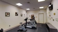 This single-floor fitness room offers a dedicated exercise space designed for cardiovascular training. The room features a drop ceiling with large recessed panel lights that provide bright, even illumination throughout the space. The walls are finished in a neutral white, accented by framed sports-themed artwork, adding a light decorative touch to the functional environment.

The flooring consists of dark rubber gym matting, providing a durable, slip-resistant surface well-suited for workout equipment. The room is equipped with several cardio machines, including a stationary upright bike, a treadmill, and an elliptical trainer, arranged to allow comfortable use of each piece of equipment. A wall-mounted panel is visible, likely serving as a control or utility interface.

Ventilation is addressed through ceiling-mounted HVAC vents, and a standard interior door provides access to the room. The overall layout is straightforward and efficient, prioritizing functionality. The space is clean, well-lit, and purpose-built for fitness use, making it a practical amenity for regular exercise routines.