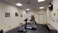 This single-floor fitness room offers a dedicated exercise space designed for cardiovascular training. The room features a drop ceiling with large recessed panel lights that provide bright, even illumination throughout the space. The walls are finished in a neutral white, accented by framed sports-themed artwork, adding a light decorative touch to the functional environment.

The flooring consists of dark rubber gym matting, providing a durable, slip-resistant surface well-suited for workout equipment. The room is equipped with several cardio machines, including a stationary upright bike, a treadmill, and an elliptical trainer, arranged to allow comfortable use of each piece of equipment. A wall-mounted panel is visible, likely serving as a control or utility interface.

Ventilation is addressed through ceiling-mounted HVAC vents, and a standard interior door provides access to the room. The overall layout is straightforward and efficient, prioritizing functionality. The space is clean, well-lit, and purpose-built for fitness use, making it a practical amenity for regular exercise routines.