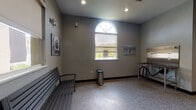 This single-floor pet grooming facility is thoughtfully designed to provide a functional and welcoming environment for pets and their owners. The space is finished with durable speckled epoxy flooring throughout, offering easy maintenance while contributing to a clean, professional aesthetic. Gray-toned walls provide a neutral backdrop, accented by dog-themed artwork and photography displayed at various points, adding personality to the utilitarian space.

The facility features multiple grooming areas equipped with professional-grade stainless steel wash tubs fitted with handheld sprayers and grooming attachments, as well as stainless steel work tables for drying and styling. A dedicated waiting area provides a comfortable bench seating arrangement beneath prominent arched windows that flood the space with natural light and offer views of the surrounding exterior. Additional arched windows appear throughout, reinforcing the bright and open atmosphere.

Recessed lighting supplements the natural light, ensuring well-lit workspaces across all grooming stations. Practical amenities include wall-mounted ventilation units, a fire extinguisher, clearly marked exit signage, and step-access features on the wash stations for ease of use. The overall design balances practicality with a welcoming ambiance suited to a professional pet care environment.