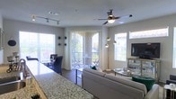 This well-appointed single-floor space is designed with an open-concept layout that effortlessly connects the kitchen, dining, and living areas into one cohesive and functional environment. The living room is anchored by a comfortable neutral-toned sofa, complemented by a sleek glass coffee table and a media console, creating a relaxed yet polished atmosphere. A ceiling fan with an integrated light fixture provides both comfort and ambient illumination overhead.

The dining area sits adjacent to the living space, furnished with a dark dining table and chairs, and is bathed in natural light streaming through large windows fitted with vertical blinds. Stylish pendant lights hang above, adding a decorative touch to the space.

One of the most notable features is the arched glass sliding door that opens directly onto a private balcony, offering views of the surrounding greenery and inviting an abundance of natural light into the interior. Additional windows along the perimeter further enhance the bright and airy feel throughout.

Underfoot, wood-style flooring runs throughout the main living areas, adding warmth and continuity to the overall design. Track lighting accents the ceiling near the kitchen transition, adding functional and aesthetic versatility. The overall ambiance is light, modern, and welcoming.