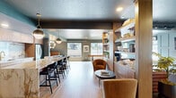Located at 245 S Orchard St in Boise, Idaho, this single-floor space showcases a carefully curated mid-century modern aesthetic that seamlessly blends warmth, texture, and functionality. The open-plan layout unifies the kitchen and living areas beneath a rich blue-gray ceiling, creating a cohesive and inviting atmosphere throughout.

The kitchen is a standout feature, anchored by a dramatic waterfall marble island with bold veining that serves as both a functional workspace and a visual centerpiece. Floor-to-ceiling walnut cabinetry lines the walls, complemented by a matching marble backsplash and a brushed gold faucet. Pendant lighting in brushed brass adds a refined glow, while light hardwood flooring runs throughout the entire space, tying each area together.

The living room is defined by a built-in wood shelving unit that acts as an elegant divider between zones, housing books, plants, and decorative objects. A fireplace with classic paneled surround anchors one wall, and a built-in window bench floods the space with natural light while offering a cozy nook with views of the surrounding greenery. A leather sofa and curved accent chairs reinforce the mid-century design language.

A built-in banquette dining nook adjacent to the kitchen adds charm and practicality. Throughout, the interplay of warm walnut wood, cool blue-gray walls, marble surfaces, and brass accents creates a polished yet livable environment.
