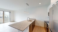 This single-floor unit showcases a contemporary design with a clean, minimalist aesthetic throughout. Light hardwood-style flooring runs continuously across the living areas, creating a cohesive and airy feel.

The kitchen is a standout feature, centered around a generous peninsula with speckled granite countertops and an integrated stainless steel sink. A sleek linear pendant light hangs above, complementing the recessed lighting overhead. White upper cabinetry reaches toward the ceiling, paired with dark lower cabinets for contrast. Stainless steel appliances — including a French door refrigerator, electric range, built-in microwave, and dishwasher — complete the well-equipped cooking space.

The open layout connects the kitchen seamlessly to the living area, which is bright and spacious. A wall-mounted mini-split unit provides climate control, while floor-to-ceiling sliding glass doors open to a private balcony, flooding the room with natural light and offering urban views of the surrounding neighborhood.

The bathroom maintains the same modern sensibility, featuring granite-topped white cabinetry, a wall-mounted toilet, and a walk-in shower with white tile surround. A warm-toned wood door with modern hardware adds a refined touch to the space's overall design.