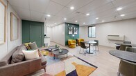 Located at 3812 Bates St in Pittsburgh's vibrant Bates Street corridor, this thoughtfully designed communal space offers a welcoming and functional environment across a single floor. The interior is defined by a striking color palette, with bold forest-green accent walls contrasting against crisp white surfaces and light wood-look flooring that runs throughout.

The space is divided into two distinct zones. A lounge area features a contemporary gray leather sofa adorned with colorful throw pillows, paired with a set of round coffee tables and anchored by a vibrant geometric area rug. Framed artwork on the white-paneled wall adds a refined, gallery-like touch. Adjacent to this is a café-style seating area furnished with dark marble-topped bistro tables and sleek green chairs, beneath a curated gallery wall of abstract paintings on the deep green accent wall.

A smaller secondary room provides a more intimate retreat, featuring a plush mustard-yellow swivel chair and a set of built-in green-painted double doors, ideal for private use or storage. Recessed lighting throughout the drop ceiling ensures bright, even illumination across all areas. A small kitchenette counter is visible along one wall, adding practical utility. Together, the spaces create a modern, inviting atmosphere well-suited for socializing, lounging, or collaborative gatherings.