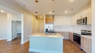 This well-appointed single-floor residence offers a thoughtfully designed layout that balances modern aesthetics with functional living. Rich hardwood-style flooring flows consistently throughout the entire unit, creating a cohesive and warm ambiance.

The kitchen serves as the centerpiece of the space, showcasing a large center island with an integrated sink and sleek light countertops, illuminated by elegant pendant lighting above. A two-tone cabinetry scheme pairs crisp white upper cabinets with natural wood-finish lower cabinets, complemented by a clean white tile backsplash. Stainless steel appliances, including a French door refrigerator with ice maker, electric range, and built-in microwave, complete the contemporary kitchen.

The living areas are bright and open, benefiting from multiple windows that invite natural light throughout. Ceiling fans add comfort to the bedrooms, each of which features neutral warm-toned walls and ample space. A walk-in closet with built-in wire shelving systems offers generous storage capacity.

The hallway connects spaces efficiently, with clean white panel doors and a sliding barn-style door adding a modern detail. The bathroom is clean and functional, featuring a shower-tub combination with brushed nickel fixtures. Overall, the space presents a clean, move-in-ready environment with modern finishes throughout.