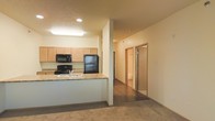 This single-floor residence is designed with an open-concept layout that seamlessly connects the kitchen and living area. The kitchen is equipped with light maple cabinetry, laminate countertops, and black appliances, including a refrigerator, electric range, and built-in microwave. A peninsula counter extends into the living space, offering additional prep space and a natural divider between the two areas. The kitchen benefits from a combination of recessed lighting and a ceiling-mounted fluorescent fixture, ensuring ample illumination throughout.

The living area features neutral-toned carpet, while the kitchen and hallway transition to hard-surface flooring, providing a practical contrast between the two zones. The textured ceiling and warm, neutral wall tones contribute to a cohesive and understated aesthetic throughout the space.

A hallway extends from the main living area, leading to additional rooms, including what appears to be a bathroom with a glass-enclosed shower and at least one other room, likely a bedroom. The layout is straightforward and functional, offering a comfortable and efficient use of space on a single floor.