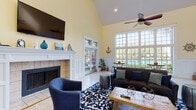 Located at 4711 S Himes Ave in Tampa, FL, this single-floor clubhouse space exudes a welcoming coastal charm with a thoughtfully curated nautical theme. The centerpiece of the main living area is a dramatic vaulted ceiling that amplifies the sense of openness, complemented by a ceiling fan and recessed lighting. Warm yellow walls and white wainscoting panels lend a clean, classic aesthetic throughout the space.

A tile-surround fireplace with a crisp white mantle serves as a focal point on one wall, with a wall-mounted flat-screen television above it. Flanking the fireplace, rich hardwood stairs with white railings add architectural interest and warmth. The living area is anchored by a bold blue-and-white patterned area rug, dark sofa, accent chairs, and a rustic wooden coffee table.

Floor-to-ceiling gridded windows bathe the space in abundant natural light while framing lush tropical landscaping and a resort-style pool beyond — a stunning visual connection to the outdoors. French doors provide direct access to the exterior.

Adjacent to the main living area, a well-appointed kitchen features white cabinetry, dark countertops, stainless steel appliances, and a breakfast bar with upholstered counter stools. A cozy bistro table positioned beneath the grand windows offers a bright, relaxing nook with picturesque pool views.