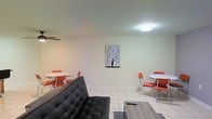 This single-floor space presents an open, combined living and dining layout designed for practical, everyday use. The main living area is anchored by a tufted black sofa, positioned facing a low-profile wooden coffee table, creating a defined seating zone. Light-colored tile flooring runs throughout, contributing to a bright and easy-to-maintain environment. The walls are finished in a soft, neutral tone that keeps the space feeling open and airy.

Two retro-style dining sets, each featuring a round white table paired with bold orange chairs with chrome accents, are positioned on either side of the room, adding a pop of color and a vintage-inspired charm to the space. A flush-mount ceiling light fixture provides overhead illumination, while a ceiling fan is partially visible, offering additional airflow. A striking black-and-white artwork depicting a figure in the rain hangs centrally on the far wall, serving as a focal point that ties the room together.

The overall ambiance is clean and straightforward, with a functional arrangement that maximizes the available floor space while keeping the interior uncluttered and inviting.