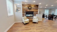 This single-floor community amenity space is thoughtfully designed to offer residents a variety of gathering and recreational options. The centerpiece of the main lounge is a striking reclaimed wood accent wall framing a sleek linear fireplace, flanked by two cream upholstered armchairs and accented by built-in display niches. Warm hardwood-style flooring flows throughout the common areas, creating a cohesive and inviting atmosphere. Natural light filters in through multiple windows fitted with blinds, brightening the neutral, softly painted walls.

Adjacent to the lounge, a secondary seating area features the same reclaimed wood fireplace wall, now paired with a wall-mounted flat-screen television, plush carpeting, and comfortable furnishings ideal for relaxing or watching a film. A welcoming entry area includes a round dining table and direct access through glass-paneled doors to the exterior.

The game room is highlighted by a full-size pool table and a large white island bar with pendant lighting in a distinctive teal hue that echoes the modern kitchenette nearby. The kitchenette is outfitted with dark cabinetry, a tile backsplash, and white countertops. A dedicated fitness center rounds out the space, equipped with treadmills, an elliptical, a multi-function weight machine, and a wall-mounted television, all set on durable rubber flooring with full-wall mirrors.
