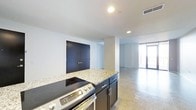 This single-floor space offers a clean, contemporary design with an open-concept layout that seamlessly connects the kitchen and living areas. The kitchen is outfitted with dark cabinetry, granite countertops, and a stainless steel range, creating a functional and stylish cooking space. A central island-style counter provides additional workspace and acts as a natural divider between the kitchen and the adjoining living area. Bold dark double doors add a striking architectural accent to the white walls, contributing to the modern industrial aesthetic. The polished concrete floors run throughout the space, lending a cohesive, urban character while reflecting the recessed ceiling lighting that keeps the interior bright and well-illuminated. Toward the far end, a large glass door or floor-to-ceiling window opens to what appears to be an outdoor balcony or terrace, allowing natural light to flood the space and offering a visual connection to the exterior. The crisp white walls, minimal detailing, and open floor plan create an airy, versatile environment well-suited for a variety of uses. The overall ambiance is modern and understated, with a thoughtful balance of functional design elements and open, uncluttered living space.