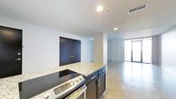 This single-floor space offers a clean, contemporary design with an open-concept layout that seamlessly connects the kitchen and living areas. The kitchen is outfitted with dark cabinetry, granite countertops, and a stainless steel range, creating a functional and stylish cooking space. A central island-style counter provides additional workspace and acts as a natural divider between the kitchen and the adjoining living area. Bold dark double doors add a striking architectural accent to the white walls, contributing to the modern industrial aesthetic. The polished concrete floors run throughout the space, lending a cohesive, urban character while reflecting the recessed ceiling lighting that keeps the interior bright and well-illuminated. Toward the far end, a large glass door or floor-to-ceiling window opens to what appears to be an outdoor balcony or terrace, allowing natural light to flood the space and offering a visual connection to the exterior. The crisp white walls, minimal detailing, and open floor plan create an airy, versatile environment well-suited for a variety of uses. The overall ambiance is modern and understated, with a thoughtful balance of functional design elements and open, uncluttered living space.