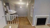Located in Arlington, Texas, this two-story home offers a well-organized layout designed for comfortable everyday living. The main living area serves as the heart of the home, featuring rich hardwood-style flooring that adds warmth and character throughout the space. A tile-surround fireplace anchors one end of the room, providing a cozy focal point, while a set of built-in wall shelves on the opposite side offers practical display and storage options. A pendant light fixture hangs above, providing focused illumination that complements the neutral gray wall tones throughout. The open layout connects the living area to a kitchen counter pass-through, allowing for easy interaction between spaces. Carpeted stairs with white railings lead to the second floor, adding a classic touch to the transition between levels. The kitchen area, partially visible from the main living space, features a countertop bar ideal for casual dining or food prep. The overall design is clean and functional, with white trim and baseboards providing crisp contrast against the gray walls. This home strikes a balance between practical design and inviting atmosphere, making it a well-suited residence in the Arlington, TX area.