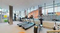 This modern fitness center is designed to provide a comprehensive workout environment within a bright, open-concept layout. The space is defined by a striking wall of floor-to-ceiling windows along one side, offering abundant natural light and views of the surrounding exterior — creating an energizing atmosphere for exercise.

The cardio zone is well-appointed with multiple treadmills, elliptical trainers, and a stationary bike, all arranged to take full advantage of the natural light and outdoor views. A dedicated free weights area includes a full dumbbell rack alongside stability balls and functional training accessories, catering to a range of strength and conditioning needs. Exposed concrete structural columns add an industrial character to the space, complementing the clean, neutral palette of grey walls and light-toned carpet flooring.

Decorative touches, including colorful artwork and a bold orange spiral motif on the floor, lend a vibrant and welcoming energy to the environment. Exposed ductwork along one section of the ceiling adds an urban, contemporary aesthetic. A staircase with metal railings connects to an adjacent level, and lockers are conveniently accessible nearby. Recessed lighting throughout ensures the space remains well-lit at all times, making this fitness center a functional and visually appealing destination for daily exercise.