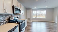 This studio unit, known as the Aspen, is located in Ann Arbor, Michigan, and offers a thoughtfully designed single-floor layout that maximizes functionality within a compact footprint. The open-concept living and kitchen area is unified by durable hardwood-style flooring that runs throughout the space, lending a cohesive and modern aesthetic.

The kitchen is a standout feature, equipped with stainless steel appliances including an electric range, over-the-range microwave, dishwasher, and refrigerator. White shaker-style cabinetry with brushed gold hardware provides ample storage, while a mosaic tile backsplash in muted grays adds visual interest. Light-toned countertops complete the clean, contemporary look.

The living area benefits from a large picture window that invites generous natural light and offers views of the surrounding neighborhood, creating an open and airy atmosphere despite the studio's scale.

The bathroom features wood-look tile flooring, a dark-toned vanity with a white countertop, and a tiled walk-in shower. A walk-in closet, visible from the main living area, provides practical storage. Recessed lighting throughout the unit ensures consistent, well-distributed illumination. The overall design palette of soft grays, whites, and warm wood tones gives the space a clean, modern character.