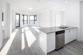This contemporary apartment showcases a thoughtfully designed open-concept layout, maximizing space and natural light. The living area is defined by expansive floor-to-ceiling windows, offering breathtaking panoramic views of the cityscape. Light wood flooring throughout adds warmth and continuity to the space.

The kitchen is a model of modern efficiency, featuring sleek white cabinetry, stainless steel appliances, and a generous island with a quartz countertop. This functional space seamlessly integrates with the living and dining areas, perfect for entertaining.

The bedroom continues the minimalist aesthetic with ample closet space and large windows. The bathroom is well-appointed with a modern vanity, large mirror, and a combination tub-shower.

Throughout the apartment, recessed lighting and strategically placed fixtures enhance the bright, airy ambiance. The neutral color palette, dominated by whites and light grays, creates a canvas for personalization.

Additional features include a dedicated closet space with wire shelving for extra storage. The apartment's design emphasizes clean lines and uncluttered spaces, embodying contemporary urban living at its finest.