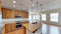 This single-story home showcases a thoughtfully designed open-concept layout that seamlessly connects the kitchen and living areas. The kitchen is a standout feature, appointed with rich honey-toned wood cabinetry that provides generous storage throughout. A curved island with a double-basin sink and light stone countertops serves as the centerpiece, offering both prep space and a natural gathering spot. Stainless steel appliances, including a gas range, wall oven, and built-in microwave, add a polished, modern touch. Pendant lighting with warm-toned glass shades hangs above the island, complementing the recessed ceiling lights that brighten the entire space. The kitchen flows effortlessly into the adjacent living area, which is bathed in natural light from a series of large windows along the exterior wall. Light wood-style flooring extends throughout, tying the open space together with warmth and continuity. The white walls and bright ceilings enhance the airy, welcoming ambiance, making the home feel open and comfortable. This well-appointed space balances practical functionality with an inviting aesthetic, ideal for everyday living and entertaining alike.