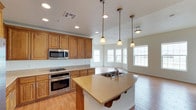 This single-story home showcases a thoughtfully designed open-concept layout that seamlessly connects the kitchen and living areas. The kitchen is a standout feature, appointed with rich honey-toned wood cabinetry that provides generous storage throughout. A curved island with a double-basin sink and light stone countertops serves as the centerpiece, offering both prep space and a natural gathering spot. Stainless steel appliances, including a gas range, wall oven, and built-in microwave, add a polished, modern touch. Pendant lighting with warm-toned glass shades hangs above the island, complementing the recessed ceiling lights that brighten the entire space. The kitchen flows effortlessly into the adjacent living area, which is bathed in natural light from a series of large windows along the exterior wall. Light wood-style flooring extends throughout, tying the open space together with warmth and continuity. The white walls and bright ceilings enhance the airy, welcoming ambiance, making the home feel open and comfortable. This well-appointed space balances practical functionality with an inviting aesthetic, ideal for everyday living and entertaining alike.