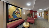 This single-floor lounge space combines contemporary design with artistic flair to create an inviting and visually stimulating environment. The room's most prominent feature is a large-scale abstract painting mounted on the wall, framed in black against warm wood paneling. The artwork's bold use of geometric shapes and vivid primary colors—yellows, blues, and reds—serves as a striking focal point that anchors the entire space.

The walls are clad in light wood paneling, adding warmth and texture, while an accent wall in deep red toward the corridor introduces a strong contrast. Wall-mounted sconce lighting complements the recessed ceiling fixtures, providing layered illumination throughout. The smooth white ceiling with flush-mount light fixtures ensures the space feels bright and well-lit.

Seating consists of low-profile, red upholstered sofas and benches, coordinating with the room's color palette. A round glass-topped coffee table with a cylindrical base anchors the seating arrangement and adds a modern touch. A lush indoor plant near the corridor brings a natural element into the space.

The patterned carpet underfoot adds visual interest and texture, while the corridor beyond hints at further rooms, suggesting this lounge functions as a shared gathering or transitional space within a larger building.