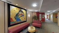 This single-floor lounge space combines contemporary design with artistic flair to create an inviting and visually stimulating environment. The room's most prominent feature is a large-scale abstract painting mounted on the wall, framed in black against warm wood paneling. The artwork's bold use of geometric shapes and vivid primary colors—yellows, blues, and reds—serves as a striking focal point that anchors the entire space.

The walls are clad in light wood paneling, adding warmth and texture, while an accent wall in deep red toward the corridor introduces a strong contrast. Wall-mounted sconce lighting complements the recessed ceiling fixtures, providing layered illumination throughout. The smooth white ceiling with flush-mount light fixtures ensures the space feels bright and well-lit.

Seating consists of low-profile, red upholstered sofas and benches, coordinating with the room's color palette. A round glass-topped coffee table with a cylindrical base anchors the seating arrangement and adds a modern touch. A lush indoor plant near the corridor brings a natural element into the space.

The patterned carpet underfoot adds visual interest and texture, while the corridor beyond hints at further rooms, suggesting this lounge functions as a shared gathering or transitional space within a larger building.