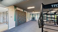 Located at 11201 Capital Blvd in Raleigh, NC, this fitness center spans a generously sized single floor divided into two distinct exercise rooms, offering a comprehensive and professionally designed workout environment. The main exercise area is outfitted with an impressive array of equipment, including treadmills, stair climbers, rowing machines, and stationary bikes, all positioned to take advantage of large floor-to-ceiling windows that flood the space with natural light and provide outdoor views. A standout feature is the full TRX suspension training rig, accompanied by battle ropes, a heavy tire, and a rock climbing wall with colorful holds — making this an ideal space for functional and high-intensity training. The ceilings are dark-painted and accented with exposed ductwork, lending an industrial character balanced by clean white walls with teal vertical stripe accents. The second exercise room offers a more open studio-style layout, complete with a mirrored wall, spin bikes with digital displays, a wall-mounted equipment rack stocked with resistance bands and medicine balls, and ample open floor space for stretching or group activity. Blue-toned commercial carpet runs throughout, and tasteful athletic-themed artwork contributes to the motivating ambiance. A small built-in storage unit and mini-fridge add a practical amenity touch.
