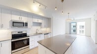 This well-designed single-floor unit offers four bedrooms, two bathrooms, a kitchen, living room, and two hallways within a efficiently organized floor plan. The interior is finished in a clean, modern aesthetic throughout, with light wood-tone flooring and crisp white walls creating a bright and cohesive atmosphere.

The kitchen serves as a central highlight, featuring white shaker-style cabinetry extending to the ceiling, a white subway tile backsplash, stainless steel appliances including a range, microwave, dishwasher, and refrigerator, and a stone-topped island that provides additional workspace and casual seating. Pendant lighting and a track lighting system illuminate the space effectively. The kitchen flows naturally into the living area, which benefits from access to an outdoor balcony framed by floor-to-ceiling sliding glass doors, inviting natural light and exterior views.

The bedrooms are clean and functional, each featuring the same light wood flooring and neutral white walls. The primary bedroom includes balcony access and a ceiling fan, while additional bedrooms offer windows with exterior views and ceiling fans. The bathrooms are finished with large-format tile flooring, white cabinetry with stone countertops, and framed mirrors, delivering a polished, contemporary look throughout.