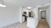 This single-floor space showcases a clean, contemporary design with a bright and open layout. The kitchen is a standout feature, outfitted with white shaker-style cabinetry that extends to the upper walls, providing generous storage. Stainless steel appliances — including a French door refrigerator, electric range, and over-the-range microwave — lend a sleek, modern aesthetic. A peninsula with a light granite countertop offers additional prep space and doubles as a casual dining or gathering area, complete with an undermount sink and a modern pull-down faucet. A subtle tile backsplash adds texture and visual interest behind the range. Light-toned hardwood-style flooring runs throughout the space, complementing the bright white walls and creating a warm, airy atmosphere. A textured ceiling adds dimension overhead, while flush-mount lighting ensures the space is well illuminated. A glimpse into an adjacent bathroom reveals consistent modern finishes, suggesting cohesive design throughout the unit. The overall ambiance is fresh, functional, and move-in ready, ideal for those seeking a well-appointed, low-maintenance living space.