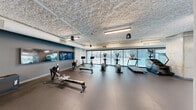 Located at 222 Third St in Detroit, MI, this fitness center spans a generous single floor and is thoughtfully divided into two dedicated exercise rooms, offering a comprehensive workout experience. The space features a modern industrial aesthetic, with textured ceilings, exposed ductwork, and concrete structural elements that lend it a clean, contemporary character.

The strength training room is equipped with a range of cable machines, weight benches, and multi-functional resistance stations finished in a bold black-and-yellow color scheme. Floor-to-ceiling glass doors and windows line the perimeter, allowing ample natural light to flood the space and providing views of the surrounding urban landscape. The room also includes a wall-mounted flat-screen display and a built-in water fountain for convenience.

The cardio room is equally well-appointed, housing a lineup of elliptical trainers, stationary bikes, and rowing machines arranged to maximize floor space and ease of movement. Large glass windows run along the exterior wall, creating an airy, open atmosphere and connecting users visually to the city outside. Additional flat-screen monitors mounted on the walls offer entertainment options during workouts.

Throughout both rooms, durable rubberized flooring provides comfort and safety, while recessed and pendant lighting ensures consistent, well-distributed illumination. The overall ambiance is clean, functional, and energizing.