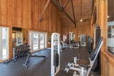 Located in Dalton, Georgia, this fitness facility is housed within a single-story structure that combines rustic architectural charm with practical workout functionality. The interior is defined by warm, tongue-and-groove wood paneling on both the walls and vaulted ceiling, supported by exposed beam framing that lends the space a distinct barn-style character and an airy, open feel.

The workout area is generously sized and laid out in an open, elongated floor plan with durable rubber flooring throughout, providing both comfort and safety during exercise. Large windows line the walls and allow abundant natural light to fill the space, offering pleasant views of the surrounding wooded landscape. French doors provide additional access points and further enhance the connection to the outdoors.

The facility is well-appointed with a comprehensive selection of equipment, including a treadmill, cable weight machines, a multi-use strength station, free weights and a dumbbell rack, resistance machines, and a flat workout bench. Wall-mounted mirrors reflect light and allow users to monitor form during exercise.

Additional amenities include a water cooler, a beverage vending machine, built-in storage cubbies, and wall-mounted accessories. A wall-mounted television adds convenience for entertainment during workouts. The overall ambiance is inviting and functional, blending a warm, lodge-inspired aesthetic with the practical demands of a fully operational fitness space.