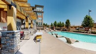 This outdoor amenity space is designed to deliver a resort-like experience, centered around a generously sized swimming pool with crystal-clear water. The pool deck is equipped with sleek, sculptural in-water lounge chairs that allow residents to relax partially submerged, while a waterfall feature adds a tranquil ambiance to the poolside environment. The surrounding concrete deck provides ample space for sunbathing and socializing, with additional seating available throughout.

Adjacent to the pool, a substantial covered pergola structure offers a shaded retreat, supported by bold stacked-stone columns and exposed heavy timber beams that lend a warm, rustic character to the space. Beneath the pergola, an outdoor entertainment area includes a wall-mounted television and comfortable seating, making it ideal for gatherings. An outdoor kitchen station with a built-in grill and stone veneer detailing further enhances the entertaining capabilities of the area.

A covered entryway with stacked-stone base accents, large grid-pane windows, and decorative outdoor lighting adds architectural refinement to the perimeter. The space is enclosed by an iron fence with mature trees lining the boundary, providing both privacy and a pleasant natural backdrop. Open skies above and abundant natural sunlight complete this vibrant, well-designed outdoor living environment.