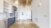This industrial-chic loft showcases a striking blend of raw architectural character and modern finishes throughout its open single-floor layout. Soaring ceilings reveal exposed concrete soffits and boldly painted orange overhead pipes and conduit, lending an authentic warehouse aesthetic to the living space. Polished concrete floors run continuously throughout, adding to the industrial ambiance while remaining easy to maintain.

The kitchen is thoughtfully appointed with two-tone cabinetry — white upper cabinets contrasted by slate-gray lower units — a classic white subway tile backsplash, and a full suite of stainless steel appliances including a French door refrigerator, electric range with microwave, and dishwasher. An open shelving section adds functional display space.

The living area is dominated by dramatic floor-to-ceiling factory-style windows framed in matte black against painted brick walls, flooding the space with natural light and offering sweeping elevated city views. The generous open layout provides excellent flexibility for furniture arrangement.

The bathroom features a clean, minimalist design with a combined shower and soaking tub enclosed by a glass panel, white walls, and a white vanity. Additional features include an electrical panel, climate control, and a walk-in closet, completing this well-equipped urban loft.