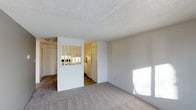 This single-floor space offers a clean, functional layout designed for comfortable everyday living. The living area is finished with wall-to-wall carpet in a neutral tone, creating a warm and inviting atmosphere. Natural light streams in through windows, brightening the room and highlighting the crisp white walls throughout. A textured popcorn ceiling adds a classic touch overhead. A defining architectural feature of this space is the partial divider wall between the living area and the kitchen, fitted with a windowed grid panel that allows light to pass between rooms while maintaining a sense of separation. This detail gives the space a distinctive character while keeping the overall feel open and connected. The kitchen is outfitted with white cabinetry and white appliances, including a full-size refrigerator, and features hard flooring that contrasts with the carpeted living space. The layout flows naturally from the main living area toward the kitchen and additional rooms beyond, making the space easy to navigate. With its neutral palette, abundant natural light, and thoughtful room division, this space provides a practical and comfortable environment suitable for a variety of uses.