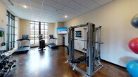 Located at 2100 Commonwealth Ave in Charlotte, NC, this well-equipped fitness center offers a comprehensive workout environment on a single floor. The space is designed with both functionality and comfort in mind, featuring polished concrete flooring with rich, earthy tones and a drop-panel ceiling with recessed lighting that provides consistent illumination throughout.

Large, floor-to-ceiling black-framed windows along one wall flood the room with natural light while offering pleasant views of the surrounding greenery, creating an energizing atmosphere for exercise. The cardio zone is anchored by multiple treadmills and an elliptical trainer, all positioned to take advantage of the natural light and outdoor views.

The strength training area is equally well-appointed, featuring a multi-station cable machine with an adjustable bench, a full rack of dumbbells, weight plates, medicine balls, and resistance bands neatly organized on wall-mounted hooks and racks. Stability balls add additional versatility to the workout options available.

A wall-mounted flat-screen television keeps users entertained during workouts, while a water fountain station provides convenient hydration. The facility also includes a dedicated entry area with a door for easy access. Overall, the space delivers a clean, modern, and fully equipped fitness experience suitable for a wide range of exercise routines.