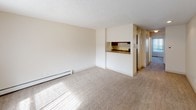 This single-floor unit offers a straightforward, functional layout designed for comfortable everyday living. The living area is generously proportioned, featuring wall-to-wall carpeting that adds warmth underfoot, a textured ceiling, and a baseboard heating unit along the wall. Natural light filters in through a large window near the private entry door, brightening the space and creating an inviting atmosphere.

The kitchen is accessible via a pass-through counter that opens toward the living area, providing a connected yet defined cooking space. It is outfitted with warm wood-toned cabinetry, granite-style laminate countertops, a stainless steel single-basin sink, a black electric coil range with overhead ventilation, a dishwasher, and a black refrigerator. The kitchen floor features hardwood-style flooring, offering a clean contrast to the cabinetry.

The bathroom continues the cohesive design with matching wood-tone cabinetry, granite-style countertops, hardwood-style flooring, and a full tub-shower combination with white tile surround. The overall color palette throughout the unit is neutral and light, with cream-toned walls that lend a clean, bright character to every room. The unit's compact, efficient footprint makes it easy to navigate and maintain.
