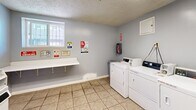 This shared laundry facility at 10216 Vultee St in Downey, California, offers a practical and efficiently organized space designed to serve its users with ease. The room is finished with light gray walls, neutral tile flooring, and a textured ceiling, creating a clean and functional environment.

The space is equipped with multiple commercial-grade front-loading and top-loading washers and dryers, accommodating several loads of laundry simultaneously. A wall-mounted folding shelf provides a convenient surface for sorting and folding clothes, while a small wall-mounted cabinet offers additional storage. Payment is supported via both coin-operated and mobile payment systems, adding flexibility for users.

A barred window along one wall allows natural light to filter into the space, enhancing the overall brightness of the room. Safety is well-addressed, with a fire extinguisher mounted on the wall and clearly posted caution and safety signage throughout. Informational notices regarding laundry room hours, best practices, and usage guidelines are displayed prominently for user reference.

A single door provides entry and exit access, keeping the layout straightforward and easy to navigate. The space is compact yet well-utilized, making it a convenient and fully equipped laundry solution for residents at this Downey location.