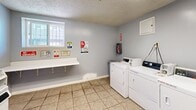 This shared laundry facility at 10216 Vultee St in Downey, California, offers a practical and efficiently organized space designed to serve its users with ease. The room is finished with light gray walls, neutral tile flooring, and a textured ceiling, creating a clean and functional environment.

The space is equipped with multiple commercial-grade front-loading and top-loading washers and dryers, accommodating several loads of laundry simultaneously. A wall-mounted folding shelf provides a convenient surface for sorting and folding clothes, while a small wall-mounted cabinet offers additional storage. Payment is supported via both coin-operated and mobile payment systems, adding flexibility for users.

A barred window along one wall allows natural light to filter into the space, enhancing the overall brightness of the room. Safety is well-addressed, with a fire extinguisher mounted on the wall and clearly posted caution and safety signage throughout. Informational notices regarding laundry room hours, best practices, and usage guidelines are displayed prominently for user reference.

A single door provides entry and exit access, keeping the layout straightforward and easy to navigate. The space is compact yet well-utilized, making it a convenient and fully equipped laundry solution for residents at this Downey location.