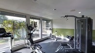 This fitness room offers a practical and well-appointed space for a variety of workout routines. The room features large floor-to-ceiling windows along one wall, flooding the interior with natural light and providing views of an outdoor pool area and surrounding greenery, creating an inviting atmosphere for exercise. The walls are finished in a two-tone scheme, with a crisp white upper half and a muted green lower half, adding a subtle visual distinction to the space. The flooring consists of dark rubber matting, a durable and practical surface well-suited for a high-traffic fitness environment. Equipment is thoughtfully arranged throughout the room and includes an elliptical trainer, a stationary spin bike, a weight bench, and a cable pulley machine mounted to the wall, catering to both cardiovascular and strength training needs. The ceiling is smooth and white, with recessed lighting and ventilation ensuring the space remains comfortable during use. Overall, the fitness room presents a clean, functional, and naturally bright environment designed to support an active lifestyle.