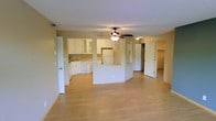 This single-floor space offers an open-concept design that seamlessly connects the living area and kitchen, creating a functional and airy environment. The light wood laminate flooring runs throughout, lending warmth and continuity across the space.

The kitchen is positioned along the back wall and features white cabinetry, including glass-front upper cabinets that add visual interest. A white refrigerator and dishwasher provide essential appliances, while a countertop peninsula serves as a natural divider between the kitchen and the living area, offering additional prep space and a casual dining option.

A ceiling fan with a built-in light fixture is centrally mounted, enhancing air circulation and providing overhead lighting to the main living space. The neutral, light beige wall tones contribute to a clean and welcoming ambiance throughout.

Toward the rear of the space, two white panel doors lead to additional rooms, one of which appears to access a walk-in closet or storage area. The layout is well-organized and practical, maximizing usable space while maintaining an open and comfortable feel suitable for everyday living.