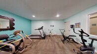 Located at 200 S Ironton St in Aurora, Colorado, this fitness room offers a dedicated exercise environment designed to support an active lifestyle. The space is finished with soft teal-painted walls that create a bright, energizing atmosphere, complemented by recessed ceiling lighting that ensures consistent illumination throughout. Durable carpet flooring covers the exercise area, providing cushioning underfoot during workouts. The room is well-equipped with a diverse selection of cardio machines, including a Spirit treadmill, multiple recumbent and upright stationary bikes, an elliptical-style stepper, and a standalone pedal exerciser. A wall-mounted mirror adds a functional touch, while decorative artwork and potted plants contribute warmth to the environment. Red lockers mounted on the wall provide convenient storage, and a paper towel dispenser and hand sanitizer station support cleanliness and hygiene. An interior window connects the gym to an adjacent area, adding a sense of openness. The room accommodates multiple users simultaneously, making it a practical and welcoming fitness amenity for the building's occupants.