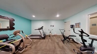 Located at 200 S Ironton St in Aurora, Colorado, this fitness room offers a dedicated exercise environment designed to support an active lifestyle. The space is finished with soft teal-painted walls that create a bright, energizing atmosphere, complemented by recessed ceiling lighting that ensures consistent illumination throughout. Durable carpet flooring covers the exercise area, providing cushioning underfoot during workouts. The room is well-equipped with a diverse selection of cardio machines, including a Spirit treadmill, multiple recumbent and upright stationary bikes, an elliptical-style stepper, and a standalone pedal exerciser. A wall-mounted mirror adds a functional touch, while decorative artwork and potted plants contribute warmth to the environment. Red lockers mounted on the wall provide convenient storage, and a paper towel dispenser and hand sanitizer station support cleanliness and hygiene. An interior window connects the gym to an adjacent area, adding a sense of openness. The room accommodates multiple users simultaneously, making it a practical and welcoming fitness amenity for the building's occupants.