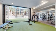 Located at 2332 N 7th St in Phoenix, AZ, this two-floor fitness center offers a comprehensive and well-appointed workout facility designed to meet a wide range of training needs. The space is thoughtfully divided across two levels, with each floor dedicated to distinct exercise functions and equipment zones.

The ground floor features a striking indoor-outdoor training area, where large folding glass walls open directly onto a generous artificial turf zone. This seamless transition between interior and exterior creates an airy, open atmosphere ideal for functional training, stretching, and free-weight exercises. Dumbbell racks, adjustable benches, and cable machines are positioned throughout, complemented by recessed lighting and views of the surrounding courtyard with its palm-lined walkways.

The upper floor is equipped with a full suite of cardio machines, including treadmills, ellipticals, and stationary bikes, all positioned along floor-to-ceiling windows that flood the space with natural light and offer views of the courtyard below. A dedicated studio area features hardwood flooring, floor-to-ceiling mirrors, a wall-mounted display screen, and medicine ball storage — ideal for group classes or individual training sessions. A bold motivational mural adds an energetic visual focal point to the strength training section, rounding out the dynamic and functional design of this modern fitness facility.