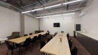 Located at 121 Westmoreland Ave in White Plains, NY, this conference room combines industrial aesthetics with modern functionality. The space features an exposed ceiling with visible ductwork and structural concrete columns, lending it a raw, urban character complemented by bright linear fluorescent lighting that ensures ample illumination throughout.

The main meeting area is furnished with long, light wood tables paired with sleek black chairs, arranged in a classroom-style layout capable of accommodating a large group. A wall-mounted flat-screen display serves as the focal point for presentations. Additional seating options include a round table with folding chairs, offering flexibility for varied meeting formats.

Along one wall, a built-in kitchenette provides dark cabinetry, a countertop with a sink, and a stainless steel refrigerator — ideal for catering needs during extended sessions. Patterned, earth-toned carpet tiles cover the floor throughout, adding warmth and acoustic absorption to the space.

A dedicated lounge nook near the glass-panel entrance features a sofa, an armchair, and a round coffee table, providing an informal setting for breakout conversations. Glass doors connect the conference room to adjacent office areas, enhancing visibility and accessibility. Overall, the space is well-suited for training sessions, team meetings, and collaborative gatherings.