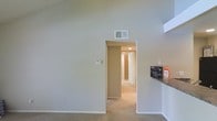 This single-floor residential space offers a clean and functional layout designed for comfortable everyday living. The interior is characterized by notably high ceilings that lend an open, airy feel to the main living area, creating a sense of spaciousness beyond what the footprint alone might suggest. Neutral, light-toned walls throughout provide a bright and welcoming atmosphere while offering a versatile backdrop for a variety of interior styles.

Soft carpeting covers the floors across the living and hallway areas, adding warmth and comfort underfoot. A central hallway extends from the main living space, providing access to additional rooms beyond, with recessed lighting overhead illuminating the passage. The hallway layout suggests a thoughtful separation between the living and private areas of the home.

A partial view of a countertop surface is visible at the edge of the living space, hinting at an adjacent kitchen or bar area. The space is equipped with ceiling-mounted HVAC vents, ensuring year-round climate control. White painted trim and baseboards provide clean, classic finishing details throughout. Overall, the space presents a straightforward and practical interior with a neutral palette, high ceilings as a standout architectural feature, and a well-connected floor plan suited to a range of occupants.