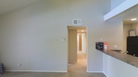 This single-floor residential space offers a clean and functional layout designed for comfortable everyday living. The interior is characterized by notably high ceilings that lend an open, airy feel to the main living area, creating a sense of spaciousness beyond what the footprint alone might suggest. Neutral, light-toned walls throughout provide a bright and welcoming atmosphere while offering a versatile backdrop for a variety of interior styles.

Soft carpeting covers the floors across the living and hallway areas, adding warmth and comfort underfoot. A central hallway extends from the main living space, providing access to additional rooms beyond, with recessed lighting overhead illuminating the passage. The hallway layout suggests a thoughtful separation between the living and private areas of the home.

A partial view of a countertop surface is visible at the edge of the living space, hinting at an adjacent kitchen or bar area. The space is equipped with ceiling-mounted HVAC vents, ensuring year-round climate control. White painted trim and baseboards provide clean, classic finishing details throughout. Overall, the space presents a straightforward and practical interior with a neutral palette, high ceilings as a standout architectural feature, and a well-connected floor plan suited to a range of occupants.