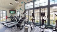 This single-level fitness center offers a comprehensive workout environment designed with both functionality and aesthetic appeal in mind. The space is defined by its striking vaulted ceilings, which soar overhead and are lined with white shiplap paneling, creating an open, airy atmosphere. Large, teal-framed floor-to-ceiling windows line the walls, flooding the interior with natural light and providing views of the surrounding outdoor landscape.

The cardio zone is well-appointed with treadmills, StairMaster steppers, elliptical trainers, and stationary cycling bikes, including Peloton-style cycles with integrated screens. A wall-mounted television provides additional entertainment during workouts. The strength training area features a multi-station cable machine, free weights, dumbbells, and weight benches, with wall mirrors expanding the sense of space.

Motivational phrases and a playful pattern of teal, green, and gold geometric triangles decorate the walls, contributing to an energetic and uplifting ambiance. The flooring consists of durable patterned carpet tiles in coordinating blue and grey tones. Pendant light fixtures hang from the peaked ceiling, complementing the natural light throughout. The overall design balances a modern, lively aesthetic with a practical, fully equipped fitness environment.