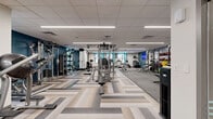 Located at 3444 S Emerson St in Englewood, Colorado, the Emerson Athletic Facility is a thoughtfully designed fitness center spanning a generous single-floor footprint. The space makes an immediate impression with its striking geometric-patterned luxury vinyl tile flooring in neutral tones of gray, beige, and cream — a design element that flows seamlessly throughout the entire facility.

The open-concept layout is divided into distinct functional zones. A dedicated cardio area lines one side of the facility, featuring a row of treadmills and stationary cycling bikes, complemented by large windows that invite natural light and provide outdoor views. Adjacent to this, a comprehensive strength training section houses a multi-station cable machine system, selectorized weight machines, and benches for a full range of resistance exercises. A free weights zone is stocked with dumbbells, medicine balls, and additional functional training equipment.

The facility is illuminated by modern linear LED lighting set against a clean drop-ceiling, creating a bright and energizing atmosphere. Bold teal accent walls punctuate the space, adding vibrancy alongside motivational wall graphics. A geometric feature wall panel serves as a striking architectural focal point near the entrance. The entry area includes a branded Emerson welcome mat, a built-in water fountain, and cubby storage for personal items, rounding out a complete and user-friendly fitness experience.