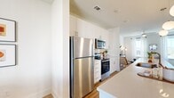 This two-story home offers a well-designed layout spread across two floors, combining modern finishes with a clean, contemporary aesthetic. Light wood-look flooring flows throughout, complementing the crisp white walls and creating a cohesive, airy ambiance.

The main floor opens into a combined living, dining, and kitchen area. The kitchen is a standout feature, boasting white shaker cabinetry extending to the ceiling, a white quartz waterfall-edge island with an undermount sink, subway tile backsplash, stainless steel appliances, and globe pendant lighting. The dining area adjoins the kitchen and offers access to a private balcony through a sliding glass door framed by sheer white curtains. The living room receives generous natural light from multiple large windows, lending a bright, open feel.

A wood-tread staircase with a metal handrail connects the two floors. Upstairs, the primary bedroom is a comfortable retreat featuring dual closets, multiple windows, and a ceiling fan. The second bedroom also enjoys ample natural light and windows on multiple walls. Both bathrooms are finished with white vanities, wood-look tile flooring, and tiled tub-shower surrounds. A hallway connects the upper-level rooms, completing this thoughtfully arranged two-story home.