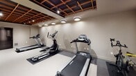 The Clinton Flats Fitness Center in Newark, NJ is a thoughtfully designed, single-floor facility that combines functional fitness amenities with contemporary architectural details. The space is defined by its distinctive open-grid ceiling, featuring a dark-framed coffered structure with warm wood tones, track lighting, and recessed flush-mount fixtures that provide even, well-distributed illumination throughout the room.

The fitness area is divided into distinct workout zones. The cardio section houses multiple Spirit-brand treadmills and elliptical trainers, positioned on dark rubber protective mats over large-format gray tile flooring. Vertical linear LED light strips accent the walls in this zone, adding a sleek, modern aesthetic. Adjacent to the cardio area is a strength training section equipped with a multi-function cable machine and an adjustable weight bench, alongside a stationary cycling bike for additional cardio options.

A separate open area features smooth, light-colored epoxy-style flooring flanked by vertical wood slat wall panels, offering a versatile space suitable for stretching, bodyweight exercises, or group fitness activities. The neutral gray walls throughout the facility contribute to a clean, focused environment. Overall, the Clinton Flats Fitness Center delivers a polished and functional workout experience with quality finishes and diverse equipment offerings.