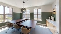 This single-floor space showcases a well-considered design that balances functionality and style across several distinct areas. A consistent design language ties the rooms together, characterized by two-tone walls with sage green on the lower half and crisp white above, complemented throughout by warm wood-tone vinyl plank flooring and recessed lighting.

The lounge area serves as a striking focal point, anchored by a floor-to-ceiling textured dark tile accent wall that lends an industrial, sophisticated edge. A sectional sofa and layered area rug create a comfortable seating arrangement, while nearby windows invite natural light and exterior views.

A dedicated workspace features a long floating white desk with warm wood cabinetry beneath, paired with open wall-mounted shelving for display and storage. Recessed lighting and wall sconces provide ample illumination for focused work.

The recreation room offers a versatile ping-pong and conference table setup beneath a decorative globe pendant light, with two large windows flooding the room with natural light. Neutral carpet and tan chairs complete the inviting atmosphere.

A well-appointed bathroom includes a sleek white quartz countertop, integrated sink, warm wood vanity cabinetry, and an elegant vertical wall sconce, finishing the space with polished, modern detail.