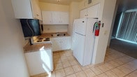 This single-floor residence offers a practical and straightforward layout with clearly defined living spaces. The kitchen is efficiently designed, featuring white upper and lower cabinetry with granite-style countertops, a white electric range, a full-size refrigerator, an over-the-range microwave, and a stainless steel sink. Light ceramic tile flooring runs throughout the kitchen and entry areas, lending a clean, easy-to-maintain surface underfoot.

The living area transitions from tile to neutral-toned carpet, creating a distinct separation between spaces. A large window fitted with vertical blinds allows natural light to fill the room, contributing to an open and airy feel. A chandelier-style light fixture near the entryway adds a subtle decorative touch.

The bedroom is generously proportioned, finished with matching carpet and windows allowing for natural light. A notable feature is the dual closet configuration, offering two side-by-side open closets with hanging rods and upper shelving—providing ample storage.

The bathroom is clean and functional, featuring tile flooring, a full-size soaking tub with a tiled surround, a shower rod, and standard fixtures. Overall, the space presents a no-frills, practical environment well-suited for everyday living.