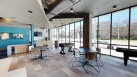 This single-story clubroom offers a well-appointed communal space designed for both relaxation and socializing. The room's most striking architectural feature is its expansive floor-to-ceiling window wall, framed in dark metal, which floods the interior with natural light and provides open views of the surrounding streetscape. The ceiling showcases an industrial aesthetic, with exposed ductwork and vent systems complemented by sleek recessed and track lighting, adding a contemporary urban character to the space.

The layout is thoughtfully organized into distinct activity zones. Multiple seating groupings, featuring modern tables paired with contemporary chairs in neutral tones, are distributed throughout, accommodating both small and larger gatherings. A foosball table serves as a central recreational focal point, adding an engaging entertainment option for residents. Toward the back of the room, a bar-height counter with seating and a mounted flat-screen television creates an inviting lounge and refreshment area. The patterned, multi-toned carpet tiles contribute visual texture and define the open floor plan, while teal accent walls add a vibrant pop of color to the overall modern, clean aesthetic.